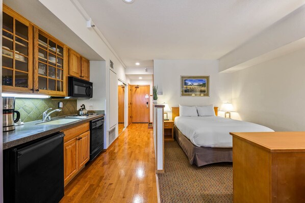 A compact hotel room with a kitchenette, a bed, and a small bedside table.