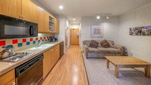 A compact kitchen with wooden cabinets, a microwave, and a kettle. There is a wooden table and a sofa in the living area.