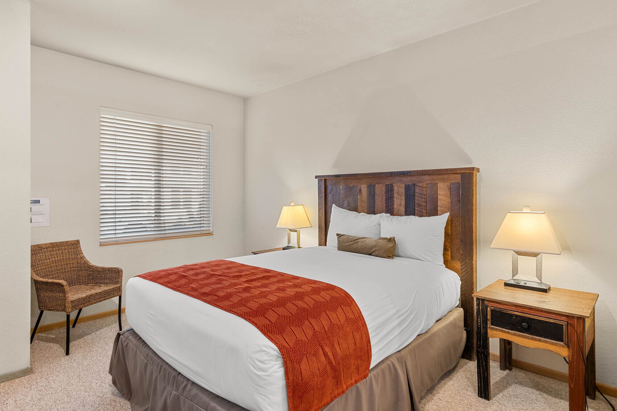 Ein Hotelzimmer mit einem großen Bett, zwei Nachttischlampen, einem Sessel und einem Fenster mit Jalousien.