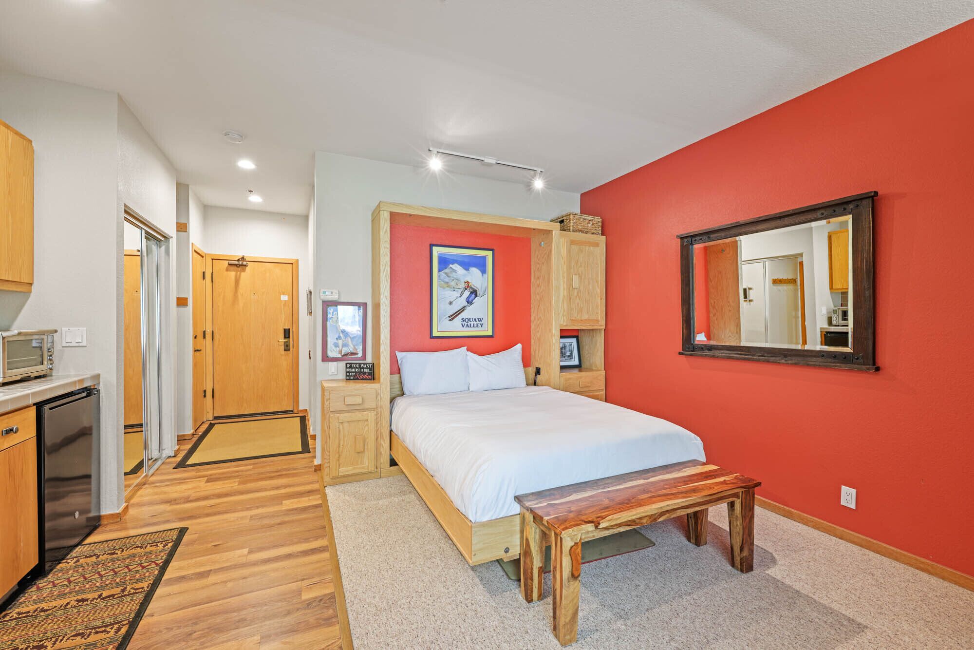 Une chambre avec un lit, un banc en bois, un mur rouge, un miroir et un coin cuisine équipé d’un four à micro-ondes.