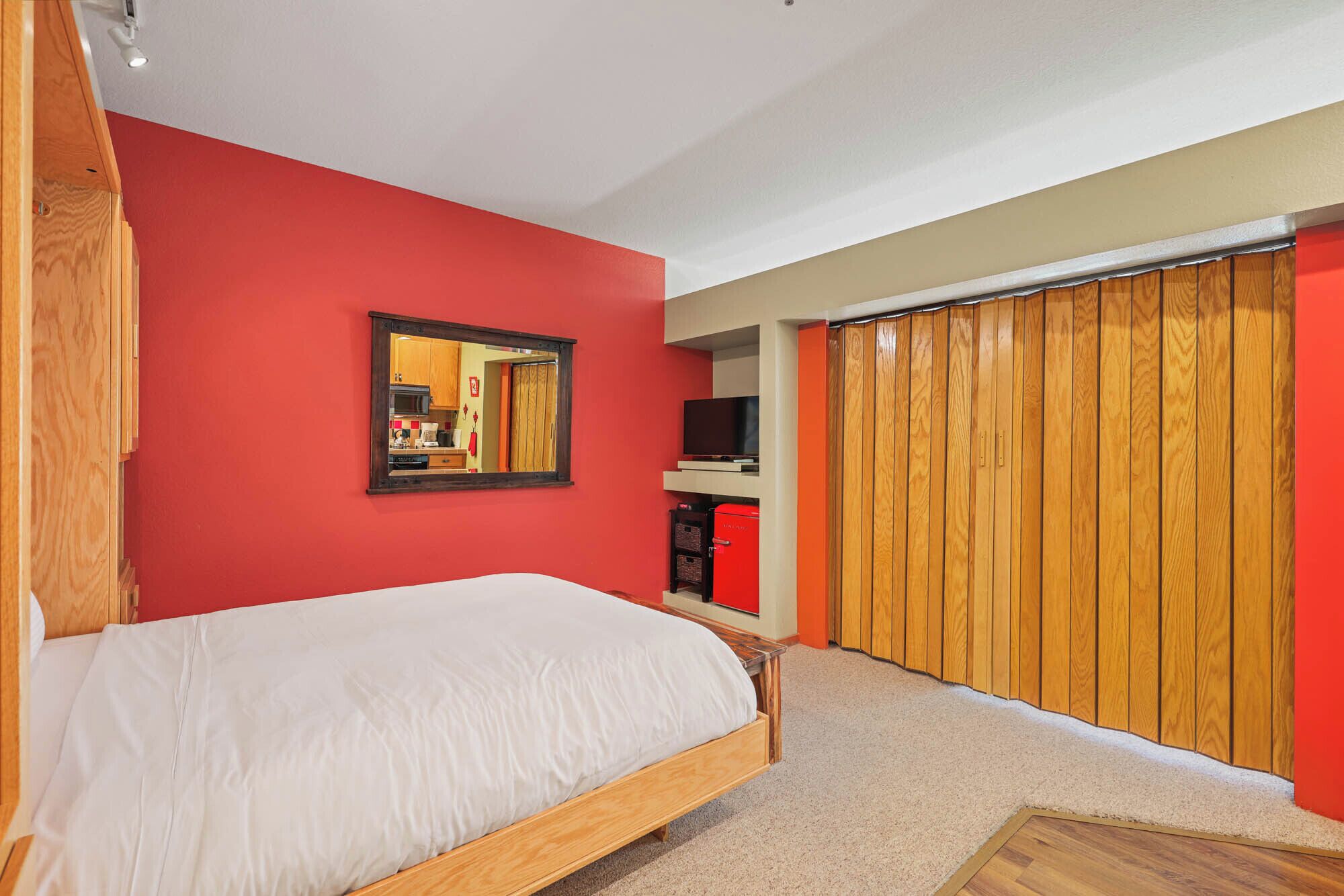 A bedroom with a wooden bed, a red wall, a mirror, a television, and a wardrobe.
