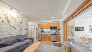 A modern living room with a grey sofa, a wooden bench, and a beige sofa with patterned cushions. The kitchen features wooden cabinets, stainless steel appliances, and a tiled backsplash. - 149 | Modern Studio w/ A/C, Wi-Fi, Views & Pool (Olympic Valley)