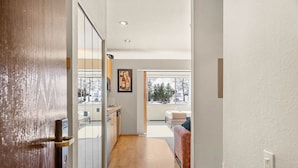 A hallway with a wooden floor, a sliding glass door, a kitchen area with a countertop and cabinets, and a large window with a view of trees and a snowy landscape. - 147 | Studio Escape - Tubs, Trails & Tranquility (Olympic Valley)