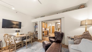 Ein modernes Wohnzimmer mit Fernseher, einer Couch, einem Essbereich mit Stühlen und einer Küche im Hintergrund.