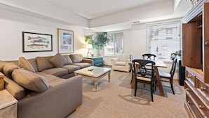 A living room with a sofa, coffee table, and dining area with chairs. There are framed pictures on the wall and a plant on the side.
