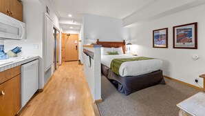 A hotel room with a bed, a wooden headboard, a small desk, and a microwave. There is a door leading to another room and a window with a view of a boat.