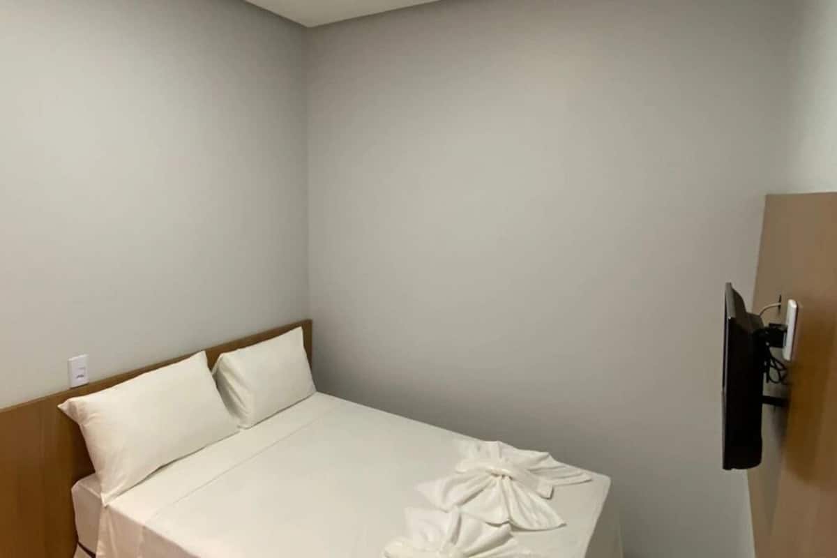 A small, neatly made bed in a corner room with a wooden headboard, a wall-mounted TV, and a power outlet on the wall.
