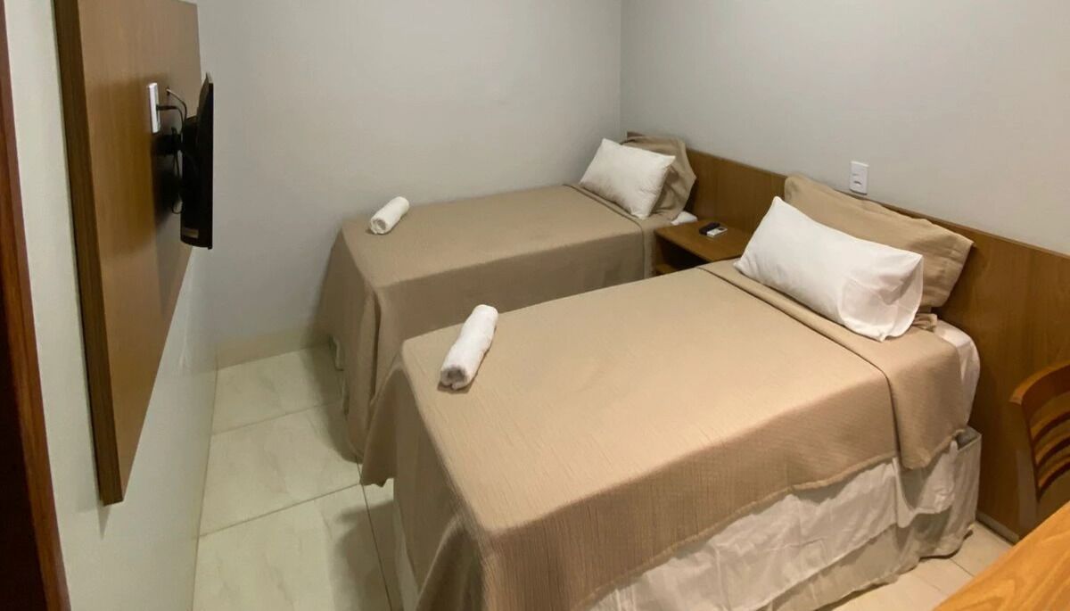 Habitación de hotel con dos camas individuales, un armario de madera, un televisor de pantalla plana montado en la pared y suelo de baldosas.