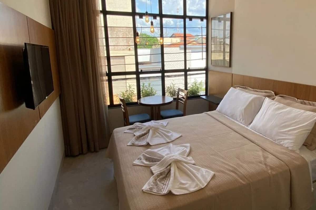 A hotel room with a bed, a TV, a window with a view of buildings, and a small table with chairs.