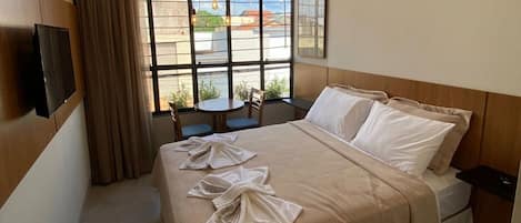 A hotel room with a bed, a TV, a window with a view of buildings, and a small table with chairs.
