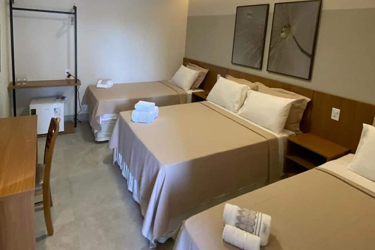 A hotel room with three single beds, a desk, a chair, and a small refrigerator.