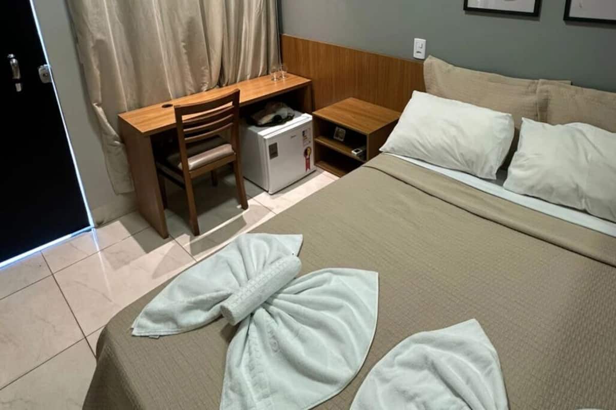 A hotel room with a bed, a desk, a chair, and a small refrigerator.