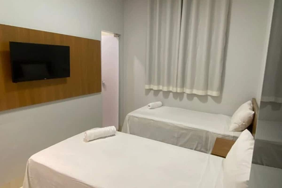 A small, clean room with two single beds, a flat-screen TV mounted on the wall, and a window with curtains.