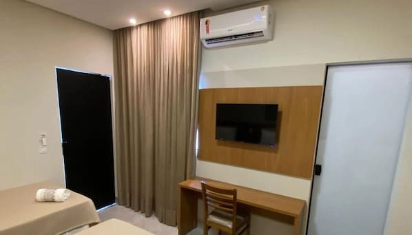Quarto de hotel com cama, escrivaninha, cadeira, televisão fixada na parede, guarda-roupa e ar condicionado.