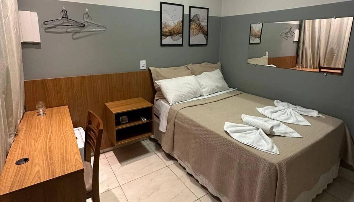 A hotel room with a bed, a wooden desk, a chair, a small shelf, and two framed artworks on the wall.