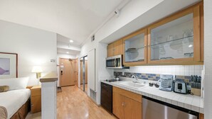 A compact hotel room with a kitchenette, a bed, a bedside table, and a lamp.