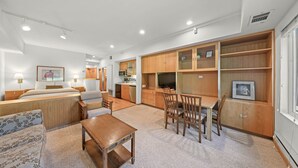 A modern hotel room with a large bed, a sofa, a small wooden table, and a dining area with chairs. There is a kitchen area with cabinets and a microwave.