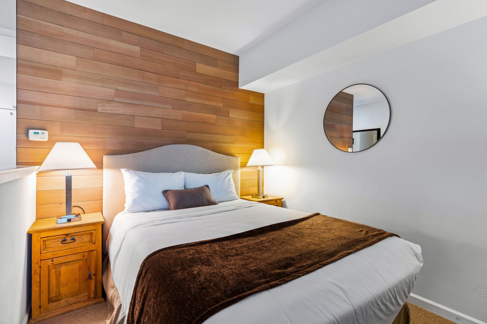Habitación de hotel impecable con cabecera de madera, un espejo redondo, dos lámparas de noche, una manta marrón y una mesita de noche de madera con cajón.