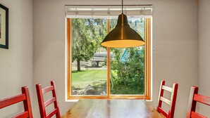 A dining area with a wooden table and red chairs, a large window with a view of trees and a building labeled "WASH SHE SHU". - 104 | Peaceful Studio - Hot Tubs + Nature Walks (Olympic Valley)