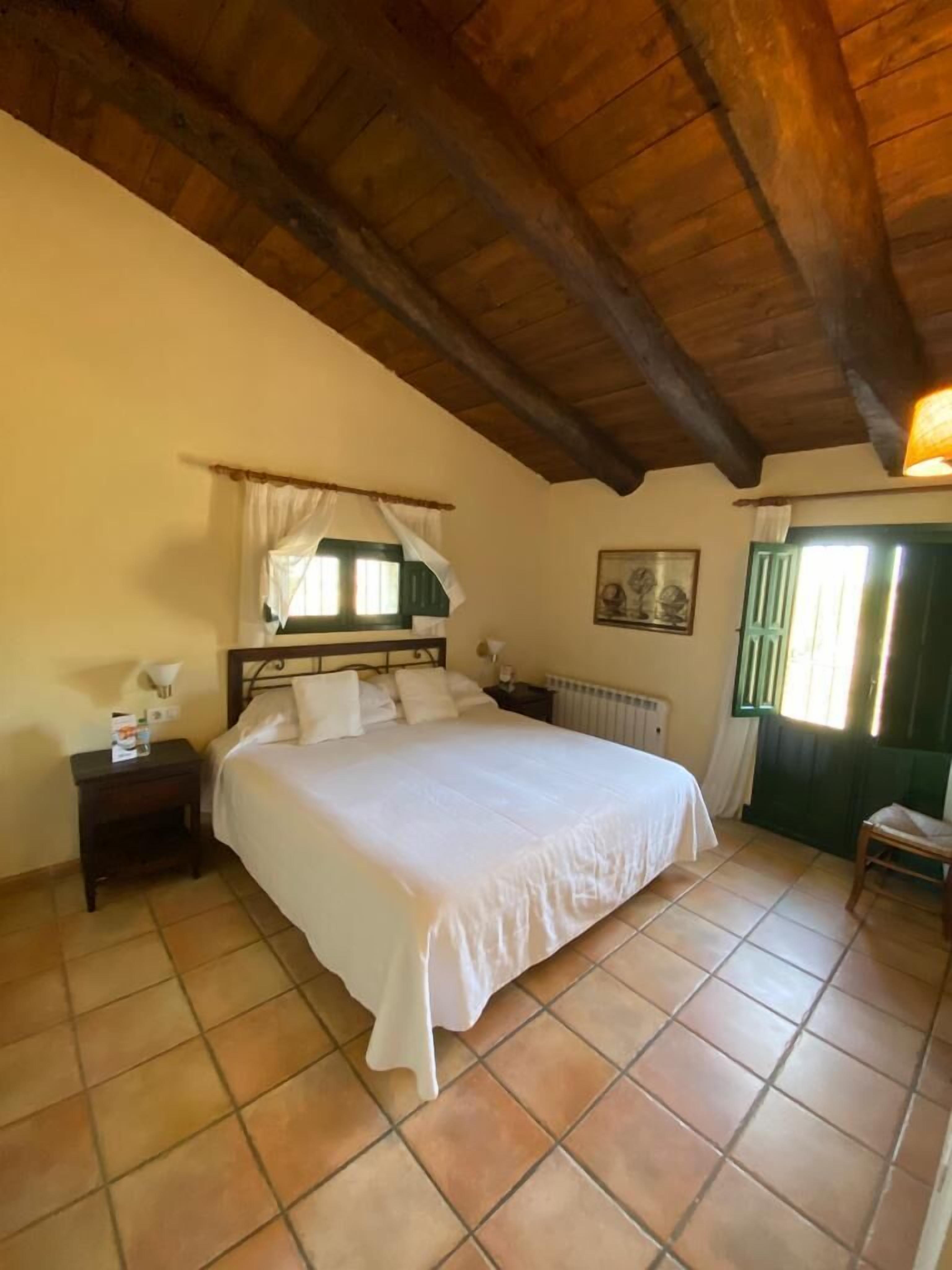 A bedroom with a wooden ceiling, a bed with white linens, a nightstand, a radiator, and a window with green shutters.