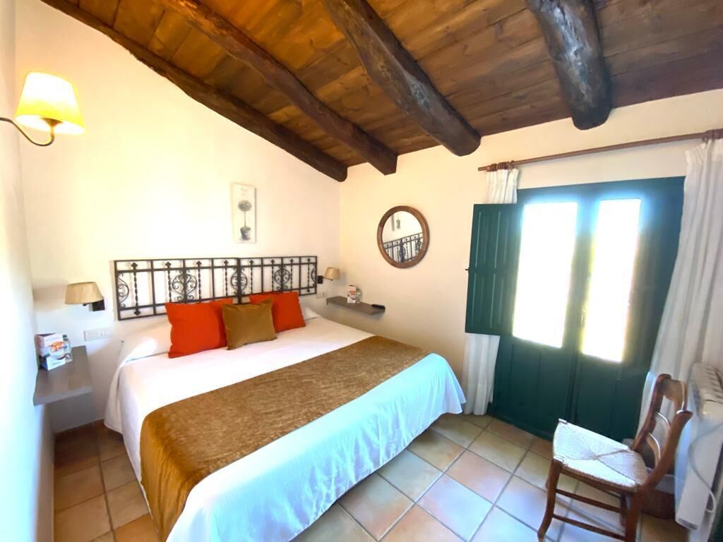 A bedroom with a bed, a chair, a nightstand, a lamp, a window with curtains, and a wooden ceiling.