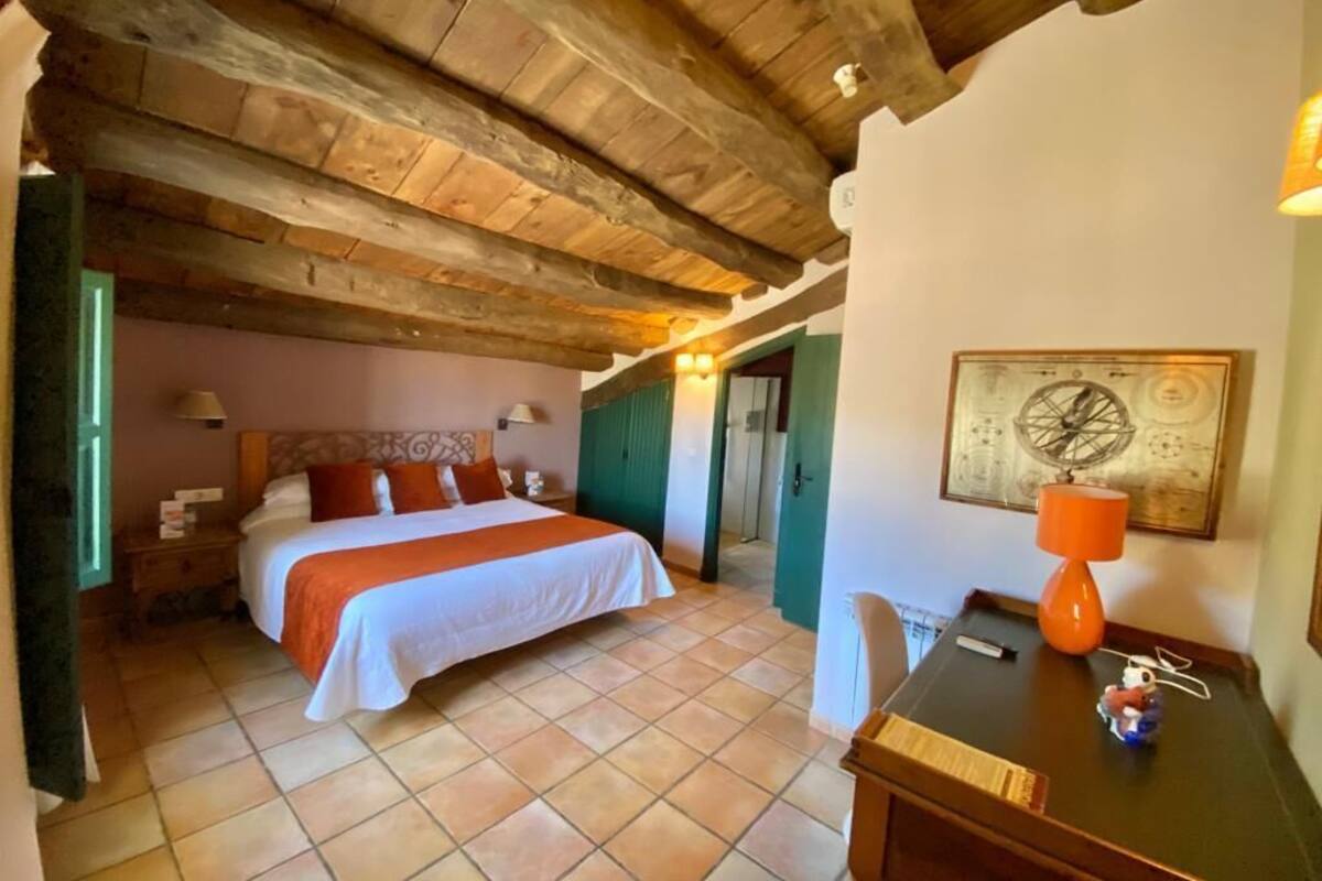A bedroom with a wooden ceiling, a bed with white and orange bedding, a desk with a lamp, and a wall with a framed picture.