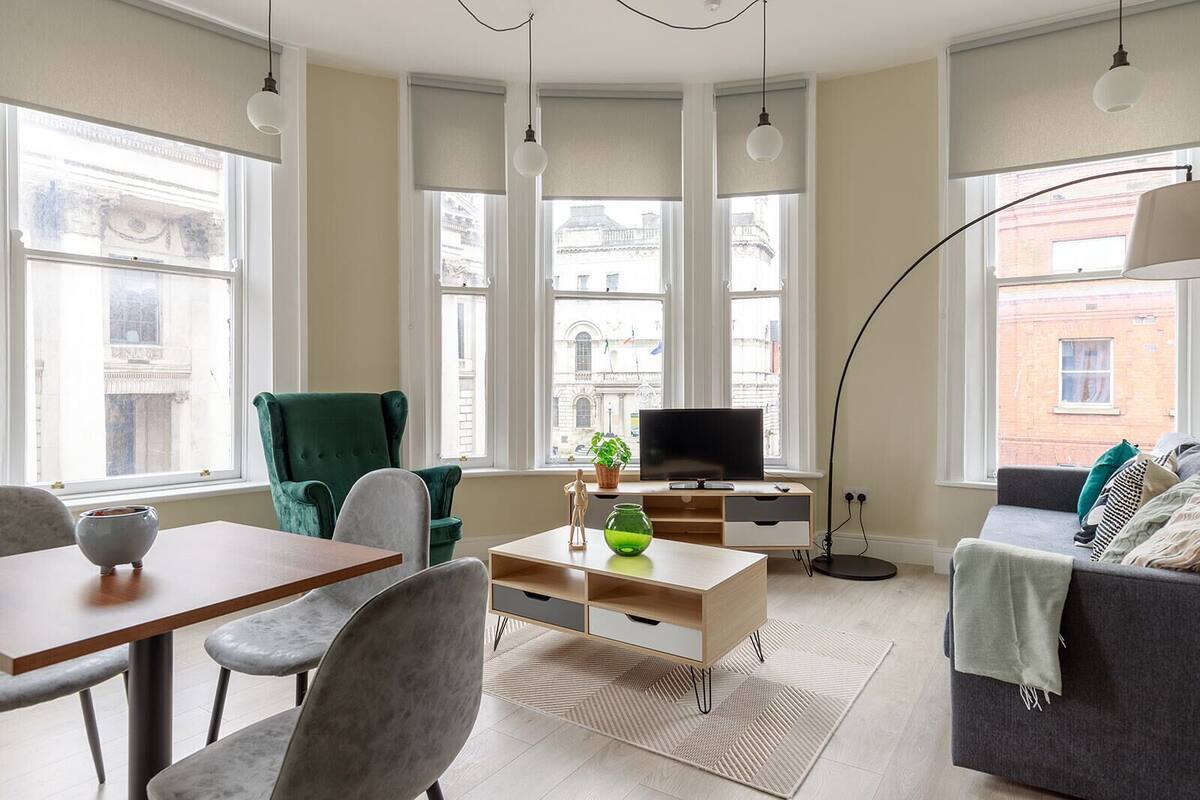 A modern living room with a sofa, a green armchair, a coffee table, and a TV. There are large windows with views of buildings outside.