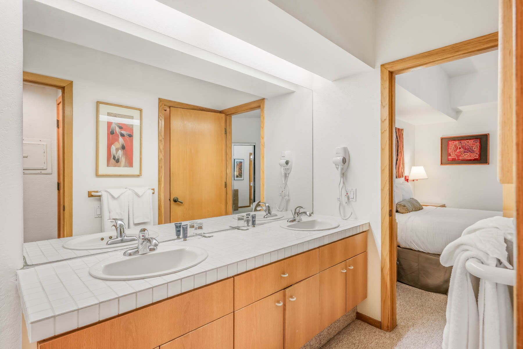 A hotel bathroom with a large mirror, two sinks, a wooden vanity, and a bed visible through an open door.