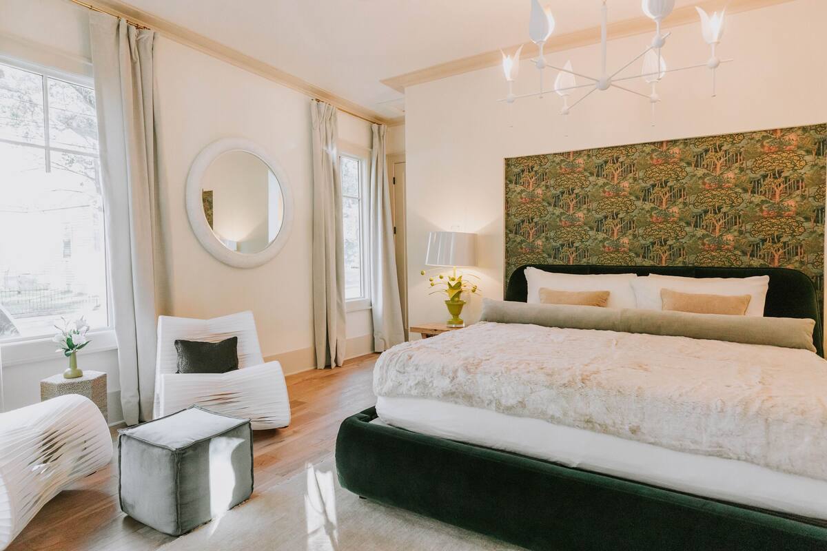 A bedroom with a large bed, a round mirror, a nightstand with a lamp, and a view of the outdoors through a window.