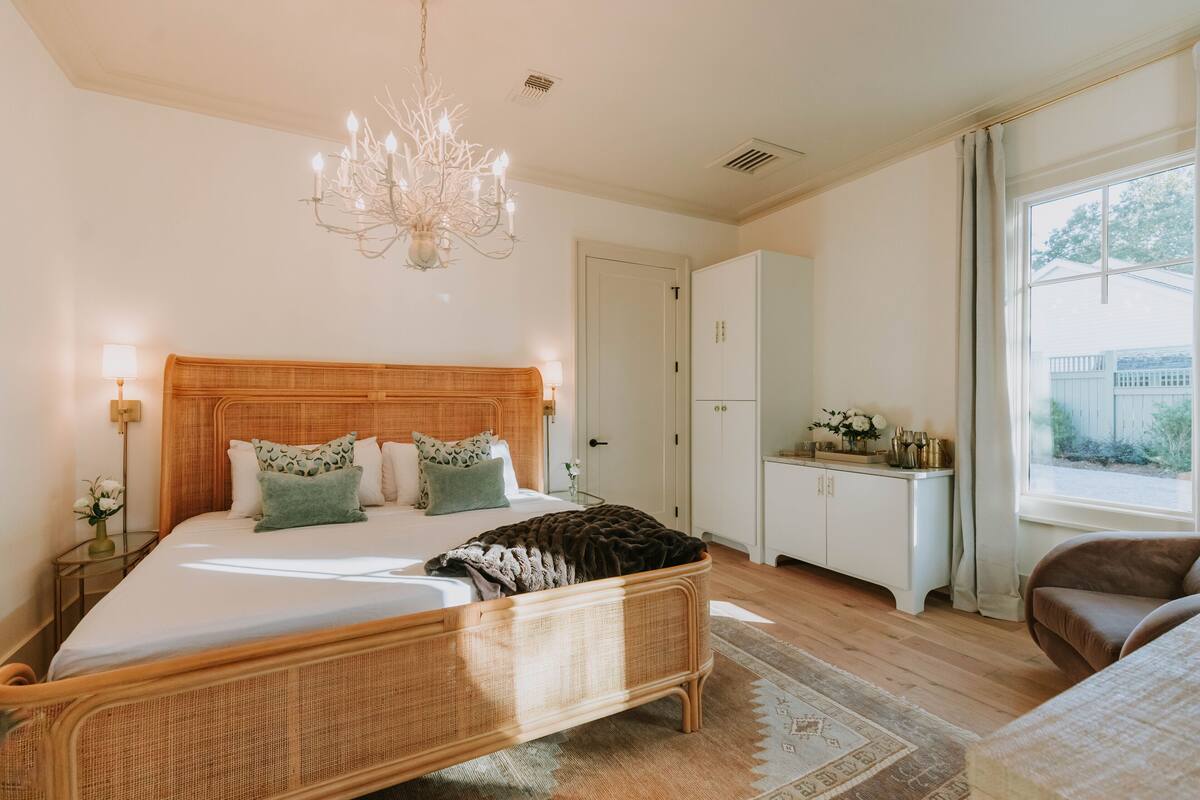 A bedroom with a large bed, a wooden headboard, a chandelier, a bedside table with a lamp, and a view of the outside through a window.