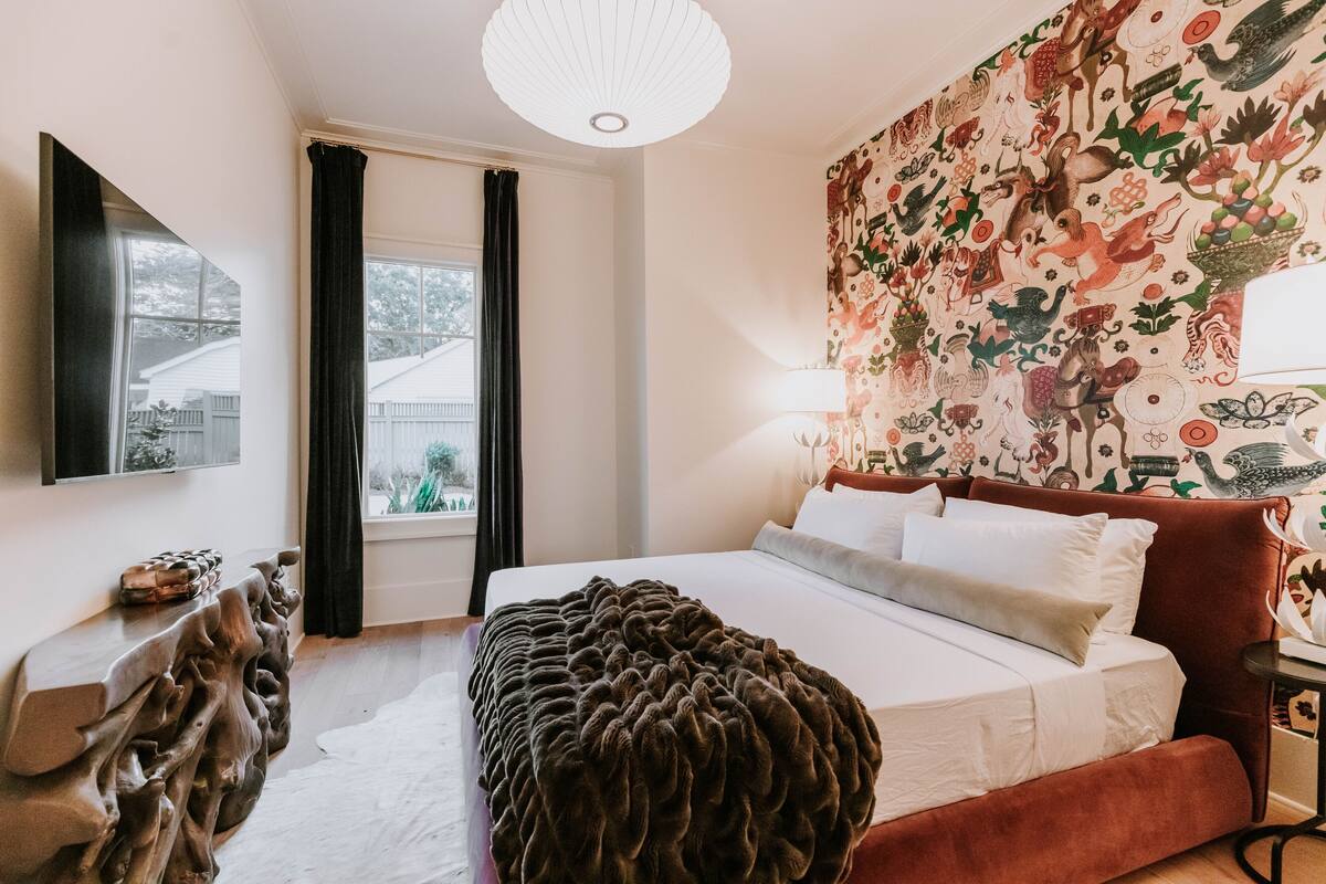 A bedroom with a large bed, a flat-screen TV, a wooden headboard, a patterned wall, and a window with curtains.