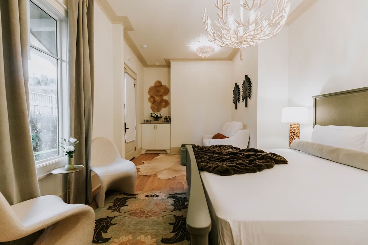 A hotel room with a bed, a chair, a small table with a vase, a chandelier, and a window with curtains.