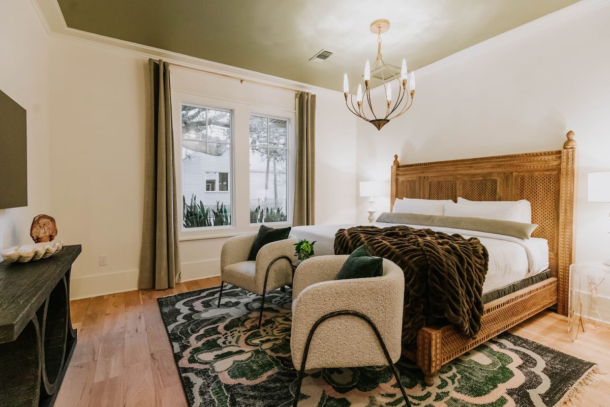 A bedroom with a large bed, two armchairs, a wooden headboard, a chandelier, and a view of trees outside.