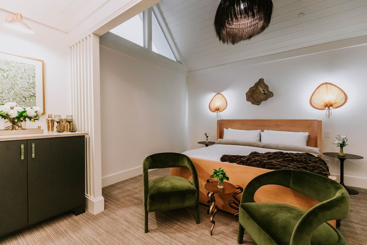 A modern hotel room with a large bed, two green chairs, a wooden side table, and a small side table with a plant.