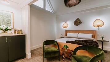 A modern hotel room with a large bed, two green chairs, a wooden side table, and a small side table with a plant.