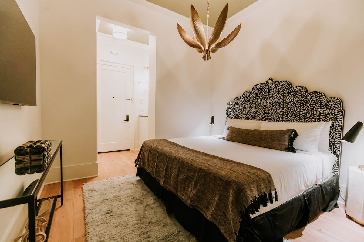 A bedroom with a bed, a hanging light fixture, a shoe rack, and a door.