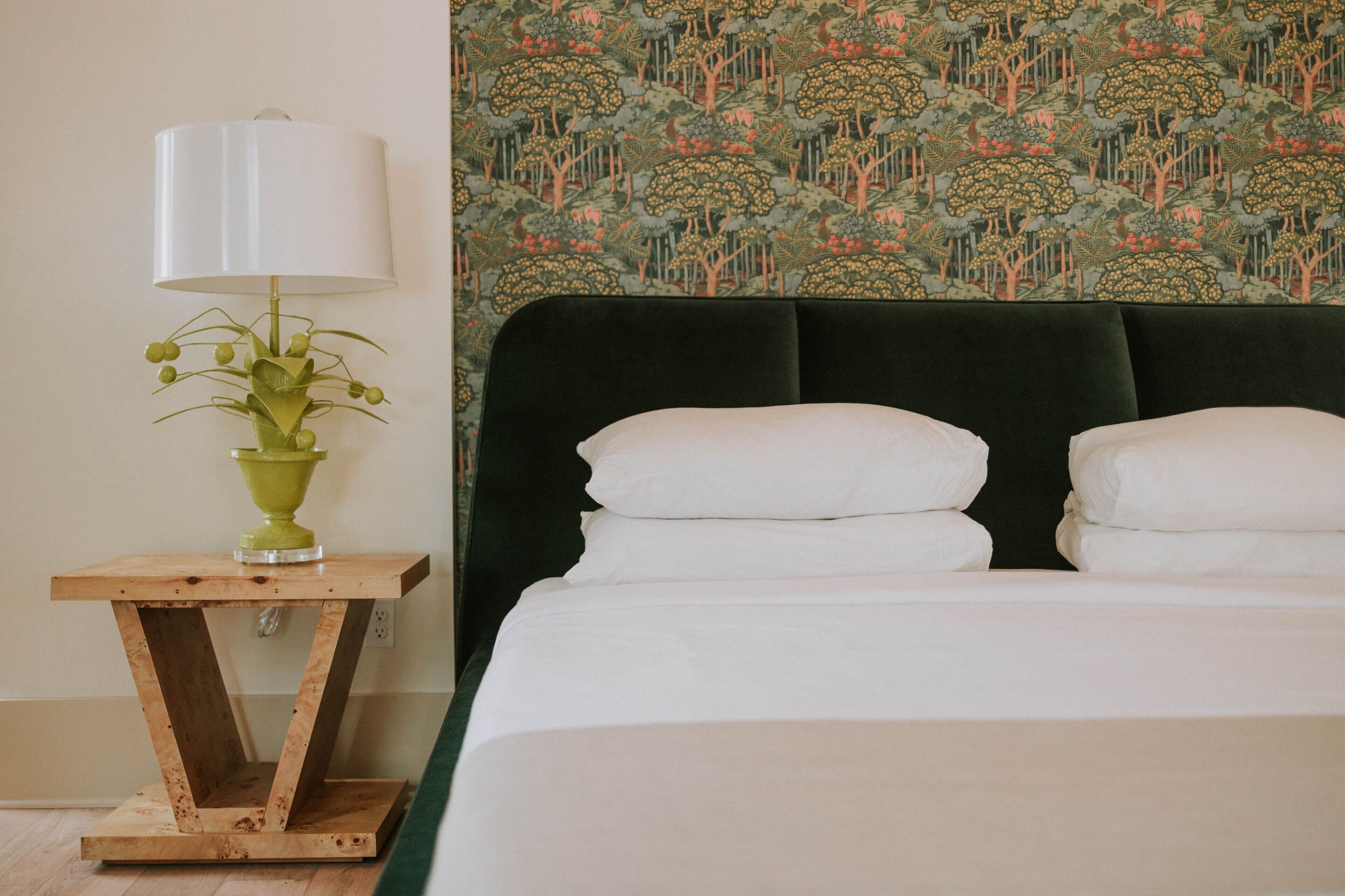 Een keurig opgemaakt bed met witte lakens, een groene vaas met bloemen en een houten nachtkastje met een lamp.