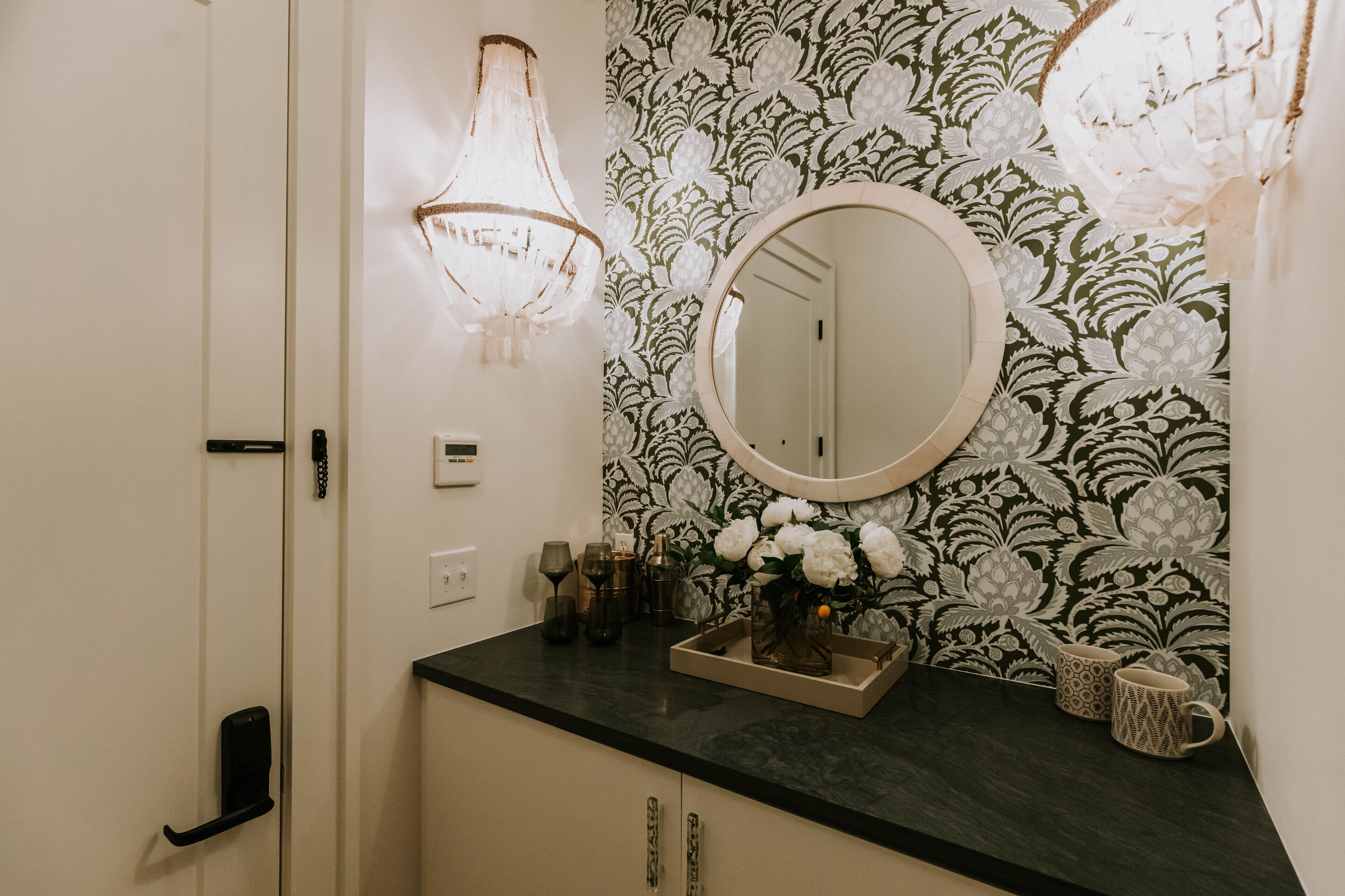 A bathroom with a black countertop, a round mirror, and a floral wallpaper.