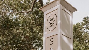 A white sign with the word "SPRINGS" and a logo, set against a backdrop of trees and a house. - The Springs Hotel (Ocean Springs)