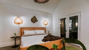 A modern hotel room with a wooden bed, two bedside lamps, a small table with a vase, and a green chair. - The Springs Hotel (Ocean Springs)