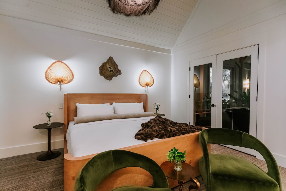 A modern hotel room with a wooden bed, two bedside lamps, a small table with a vase, and a green chair.