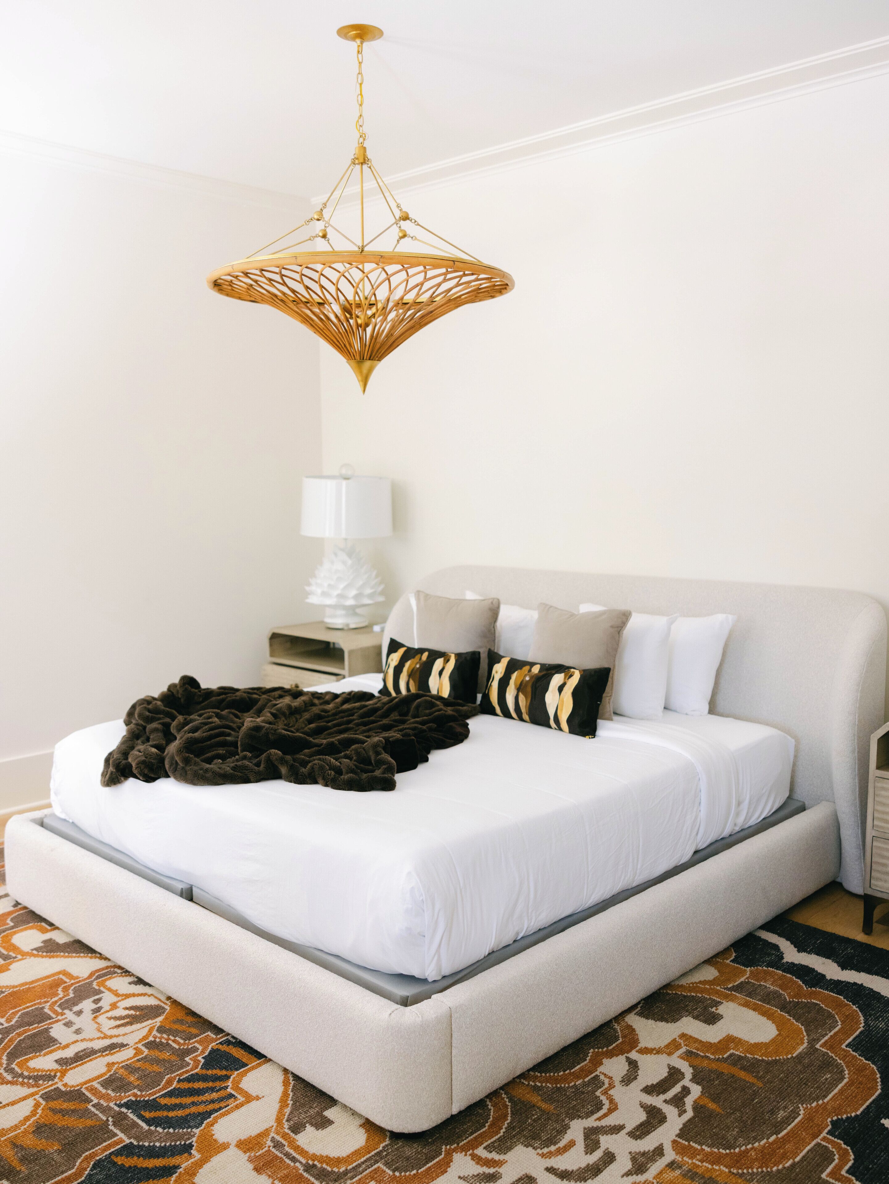 Una cama bien hecha con colcha blanca y manta oscura, cabecera tapizada en tono beige, alfombra estampada y una lámpara de araña dorada colgante.