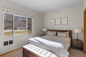 A bedroom with a large bed, two bedside tables, a bench, a window with a view of a river and hills, and three framed pictures on the wall. - Slopeview End | End-Unit King 1BD, Views, 159 (Olympic Valley)