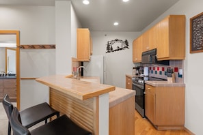 A compact kitchen with wooden cabinets, a built-in microwave, and a small island with seating. - Slopeview End | End-Unit King 1BD, Views, 159 (Olympic Valley)