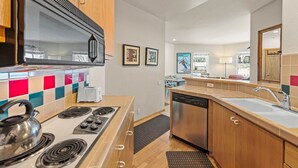 A compact kitchen with a stove, microwave, and dishwasher. There are wooden cabinets, a tiled backsplash, and a window with a view.
