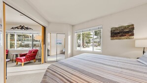 A spacious bedroom with a large bed, a red chair, a small table, and a view of the outdoors through a sliding door.