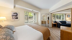 Quarto com uma cama grande, um sofá e uma escrivaninha. Há janelas com persianas, um abajur e um quadro na parede.