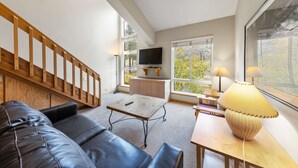 Una sala moderna con una escalera de madera, un televisor de pantalla plana, una mesa de comedor y un ventanal con vistas a árboles.