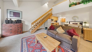 Un salón amplio con una escalera de madera, un conjunto de sofás, una mesa de centro y una cocina al fondo.