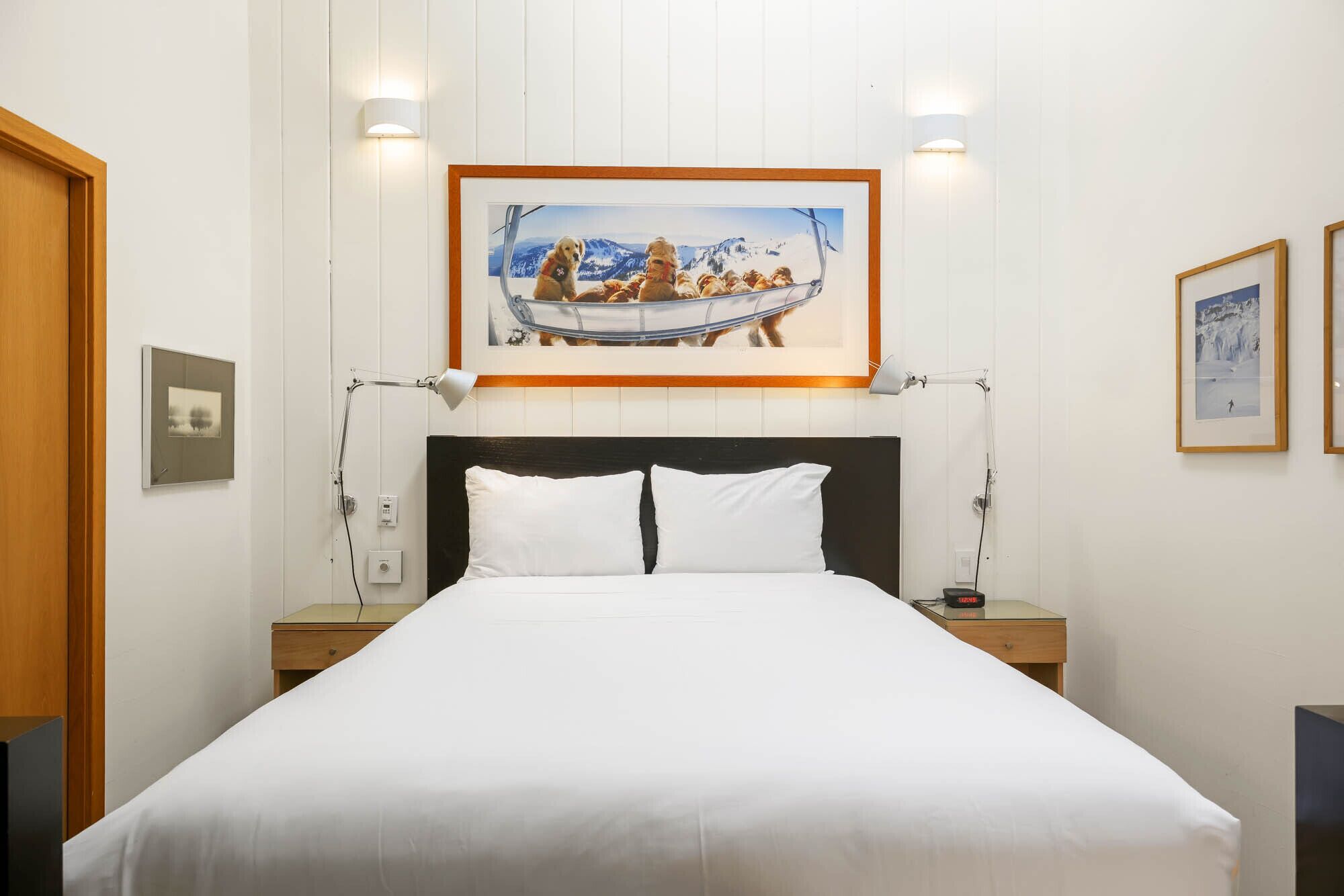Une chambre d’hôtel avec un grand lit, deux tables de chevet, un tableau représentant un chien sur un bateau, accroché au mur, et une lampe fixée au mur.
